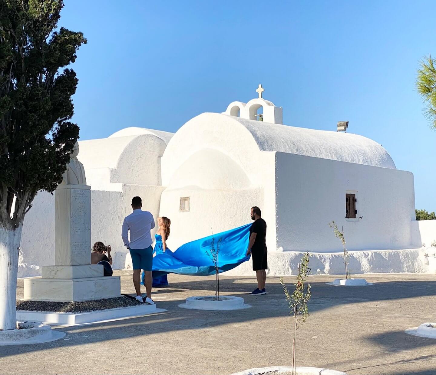 Woman in a blue flying dress poses in front of a church while an assistant hold up her dress tail